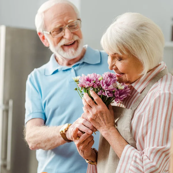 Selective Focus Senior Woman Apron Smelling Bouquet Smiling Husband Kitchen — Stock Photo, Image