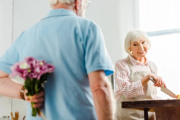 Selective Focus Smiling Senior Woman Looking Husband Bouquet While Cooking — 스톡 사진