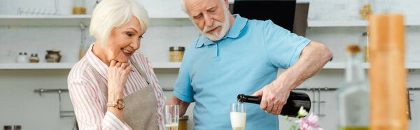 Panoramic shot of smiling woman looking at husband pouring champagne in kitchen
