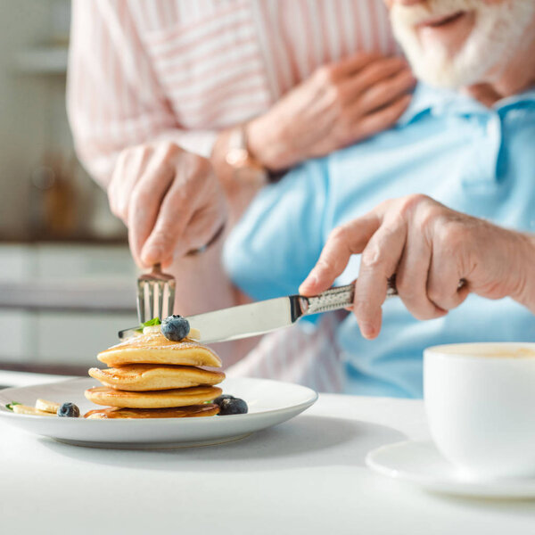 Cropped view of man cutting pancakes with blueberries by wife in kitchen
