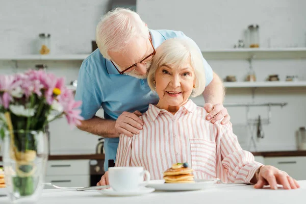 年长男人有选择性地把焦点放在抱着妻子 用煎饼和咖啡看着镜头的桌上 — 图库照片