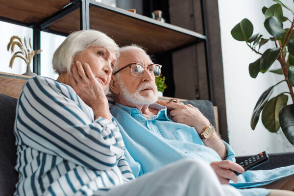 Low angle view of serious senior couple watching tv on sofa at home