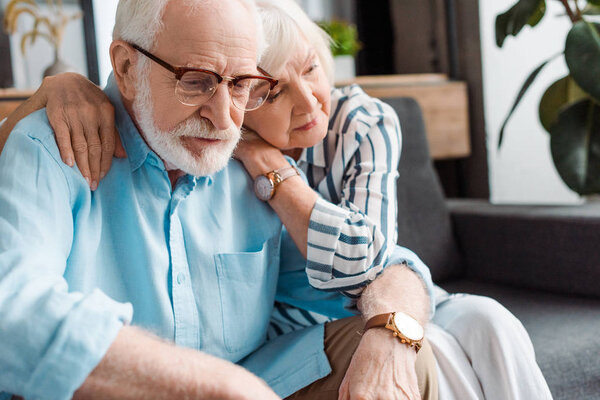Senior woman hugging sad husband on couch at home