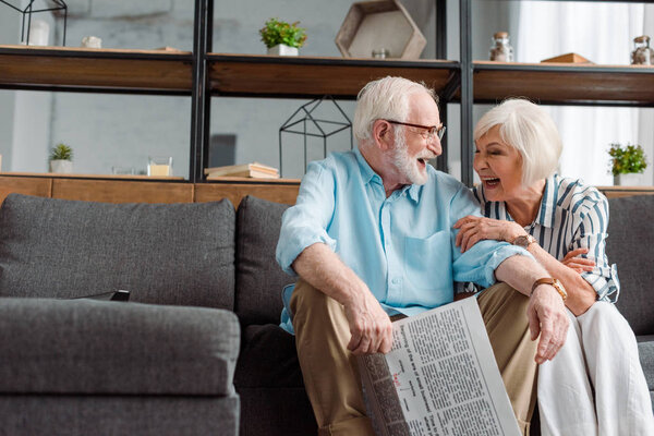 Senior couple with newspaper laughing while sitting on couch on living room