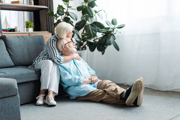 Starsza Kobieta Obejmująca Męża Podłodze Odwracając Wzrok Salonie — Zdjęcie stockowe