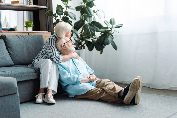 Senior Woman Embracing Husband Floor Looking Away Living Room — Stock Photo, Image