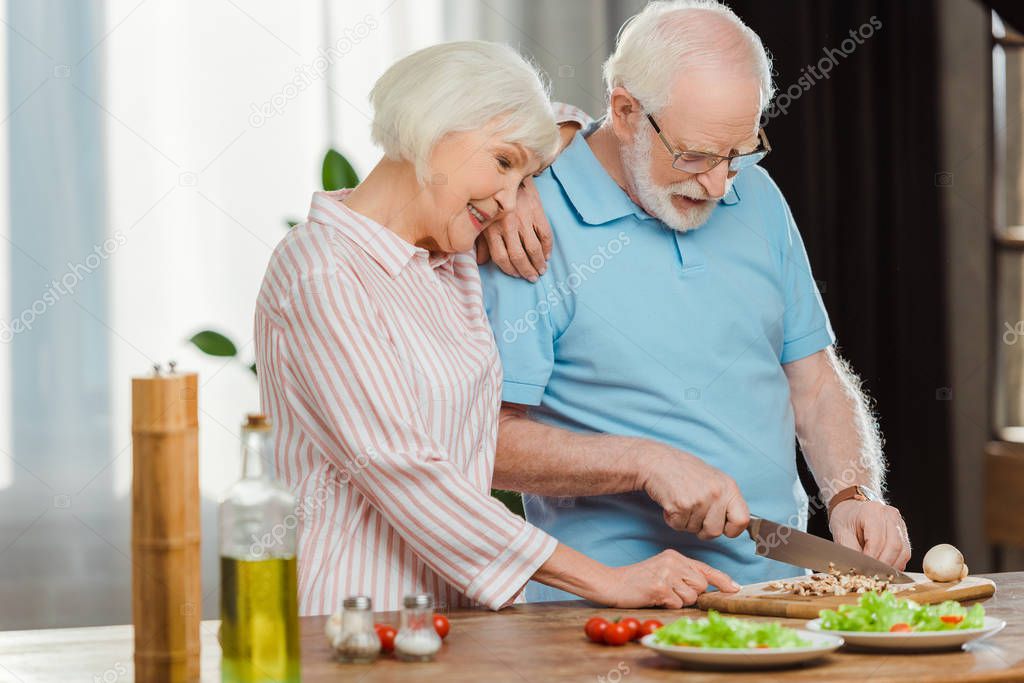Smiling senior woman standing by husband cutting vegetables on kitchen table 