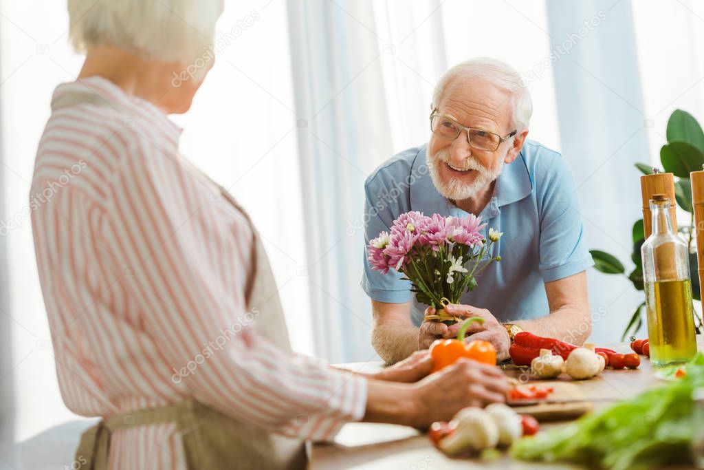 Selective focus of smiling senior man with bouquet looking at woman cooking on kitchen table