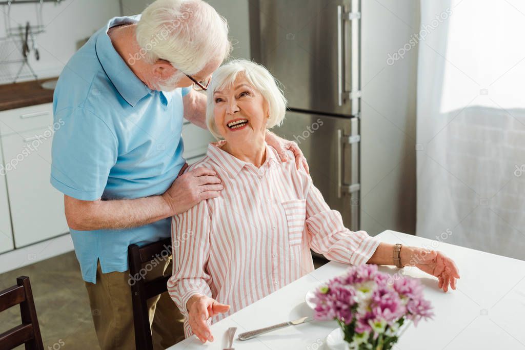 Selective focus of senior man embracing smiling wife at table in kitchen