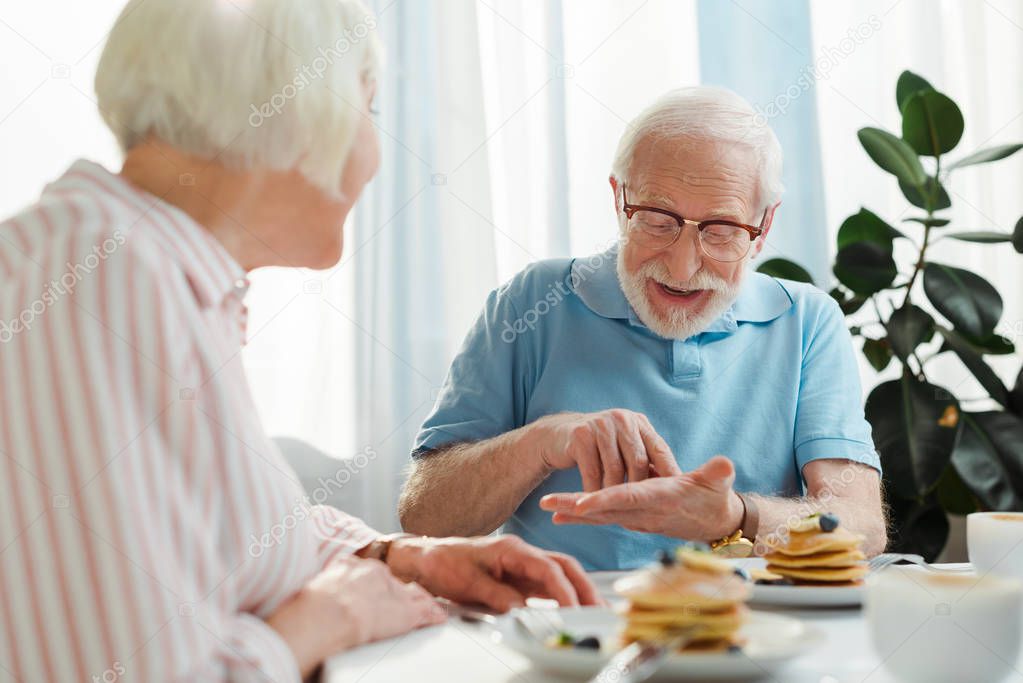Selective focus of senior man talking to wife by coffee and pancakes on table 