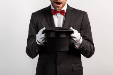 cropped view of shocked magician holding hat, isolated on grey clipart