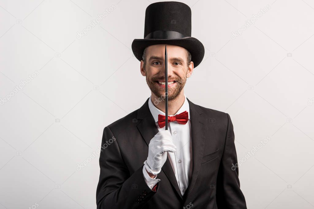 cheerful magician in suit and hat holding wand, isolated on grey