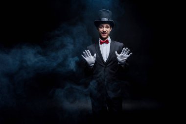 smiling magician in suit and hat gesturing in dark smoky room clipart