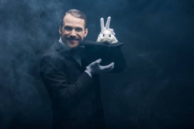 Takım elbiseli pozitif sihirbaz şapkalı beyaz tavşanla numara yapıyor, dumanlı karanlık oda