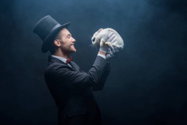 Gülümseyen sihirbaz takım elbiseli ve şapkalı beyaz tavşancık, dumanlı karanlık oda