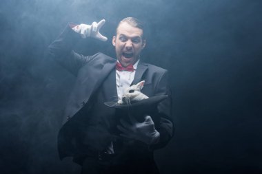 duygusal sihirbaz bağırıyor, işaret ediyor ve beyaz tavşanı şapkada tutuyor, dumanlı karanlık oda