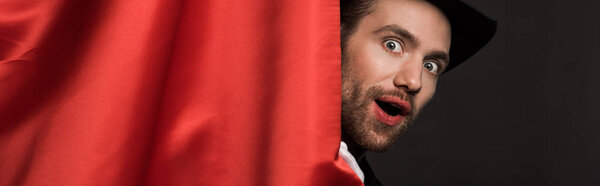 panoramic shot of shocked handsome magician in suit and hat in circus with red curtains