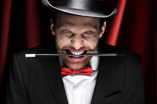professional magician in suit and hat holding wand in teeth in circus with red curtains
