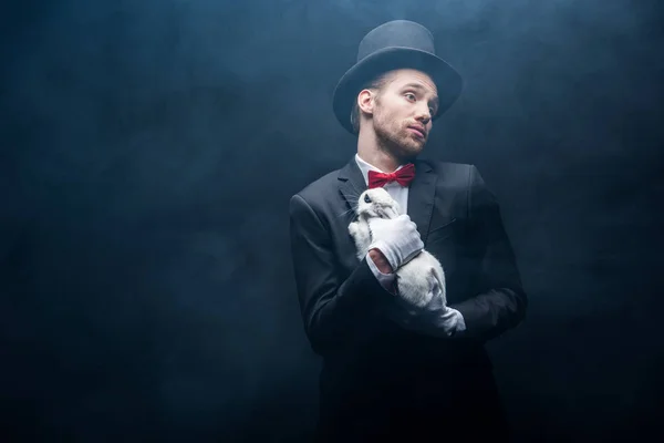 スーツを着た混乱した魔術師と白いウサギを抱えた帽子煙の暗い部屋 — ストック写真