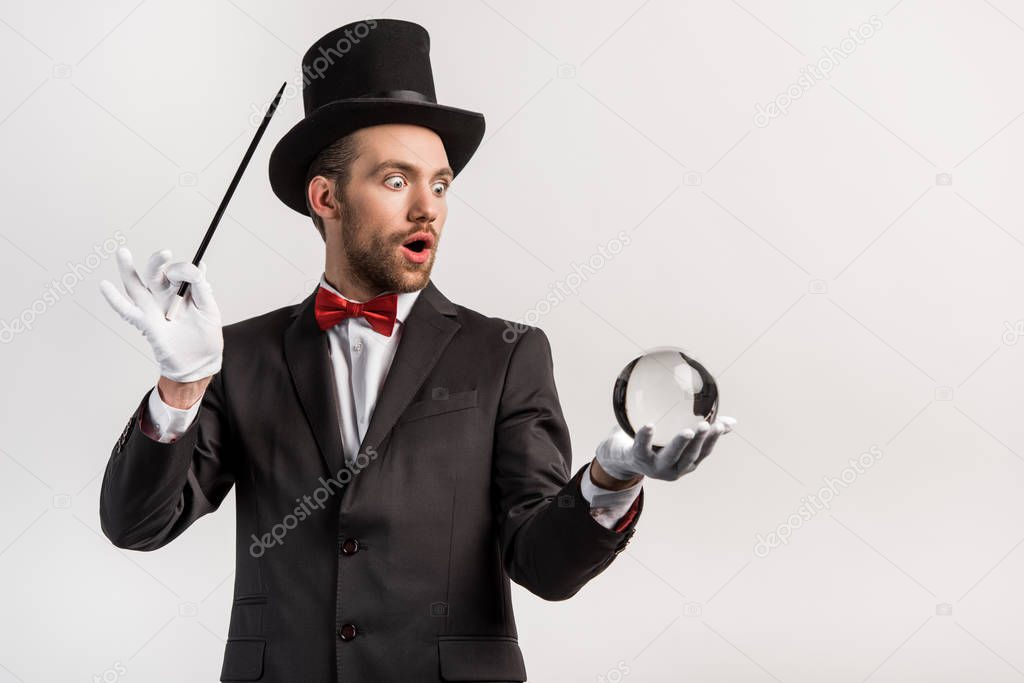 shocked magician holding wand and magic ball, isolated on grey