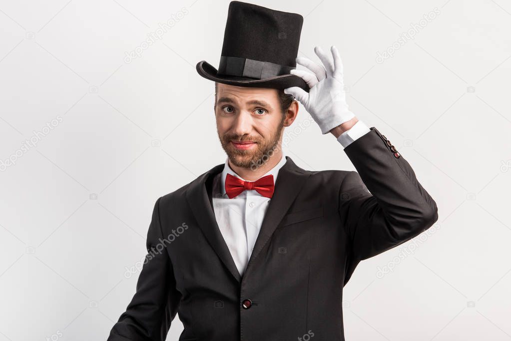 handsome happy magician adjusting hat, isolated on grey