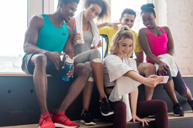 Gülümseyen çok kültürlü zumba dansçıları dans stüdyosundaki bankta dinlenirken selfie çekiyorlar.
