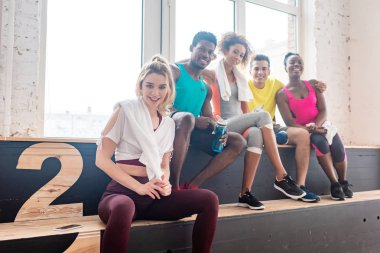 Dans stüdyosundaki bankta kameraya gülümseyen çok kültürlü zumba dansçılarının seçici odağı