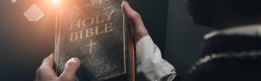 partial view of catholic priest holding holy bible near confessional grille in dark with rays of light, panoramic shot clipart