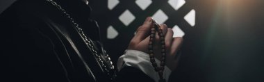 partial view of catholic priest holding wooden rosary beads near confessional grille in dark with rays of light, panoramic shot clipart