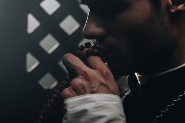 young catholic priest kissing wooden rosary beads near confessional grille in dark with rays of light clipart