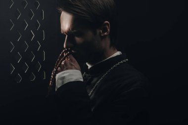 young thoughtful catholic priest kissing wooden rosary beads near confessional grille in dark with rays of light clipart