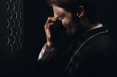 young tense catholic priest touching face with closed eyes near confessional grille in dark with rays of light