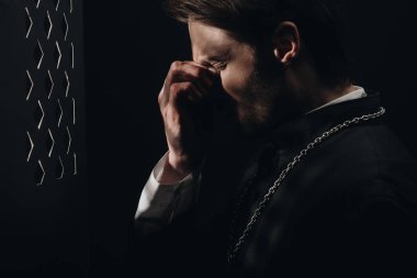 young tense catholic priest touching face with closed eyes near confessional grille in dark with rays of light clipart