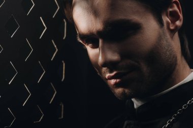portrait of young tense catholic priest looking at camera near confessional grille in dark with rays of light