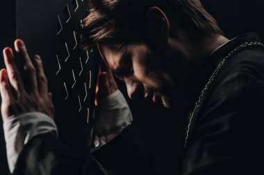 young tense catholic priest with closed eyes leaning on confessional grille in dark with rays of light