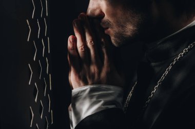 cropped view of catholic priest praying near confessional grille in dark with rays of light clipart