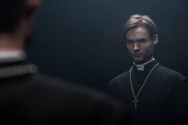 young tense catholic priest looking at own reflection isolated on black