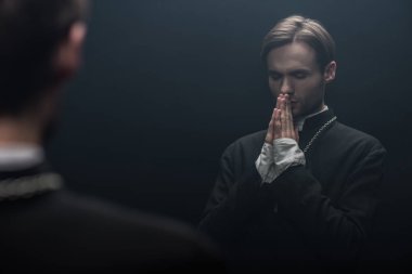 young serious catholic priest praying with closed eyes near own reflection isolated on black