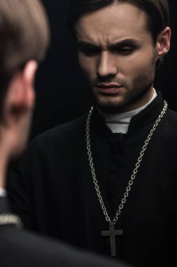 young serious catholic priest thinking while standing near own reflection isolated on black clipart
