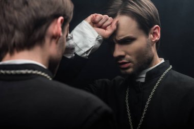young tense catholic priest holding fist near forehead while standing near own reflection isolated on black clipart