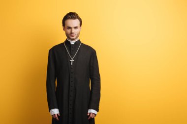 young, confident catholic priest looking at camera while standing isolated on yellow clipart