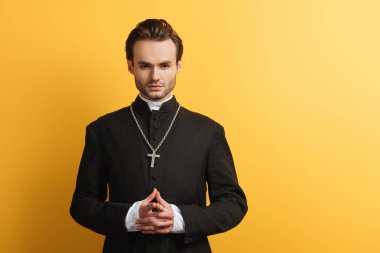 serious catholic priest standing with clenched hands while looking at camera isolated on yellow