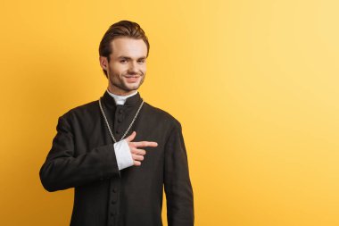 smiling catholic priest looking at camera and pointing with finger isolated on yellow