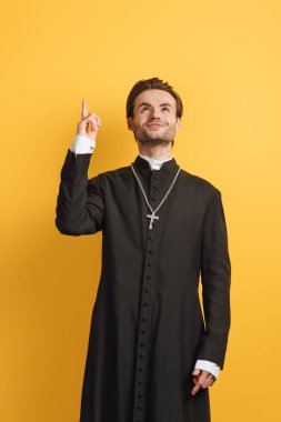 smiling catholic priest looking up and pointing with finger isolated on yellow clipart