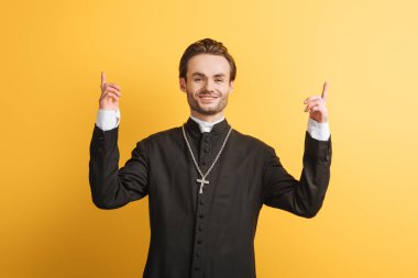 smiling catholic priest pointing up with fingers while looking at camera isolated on yellow