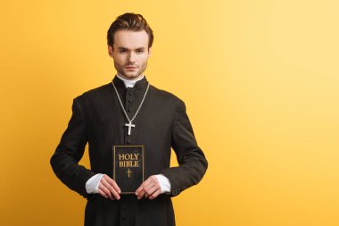 young, confident catholic priest looking at camera while holding bible isolated on yellow