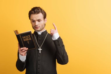 thoughtful catholic priest showing idea sign while reading bible isolated on yellow
