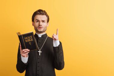 thoughtful catholic priest looking up and showing idea sign while holding open bible isolated on yellow