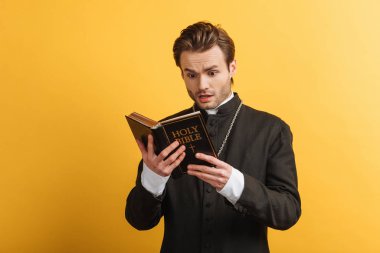 shocked catholic priest reading holy bible isolated on yellow