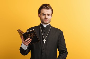 surprised catholic priest looking at camera while holding bible isolated on yellow